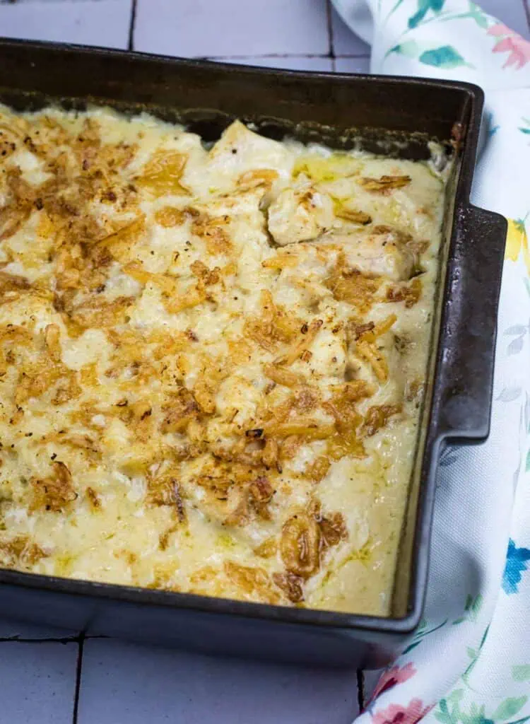 A rectangular baking dish filled with a creamy, baked casserole topped with crispy, browned onions sits on a tile surface next to a patterned cloth.