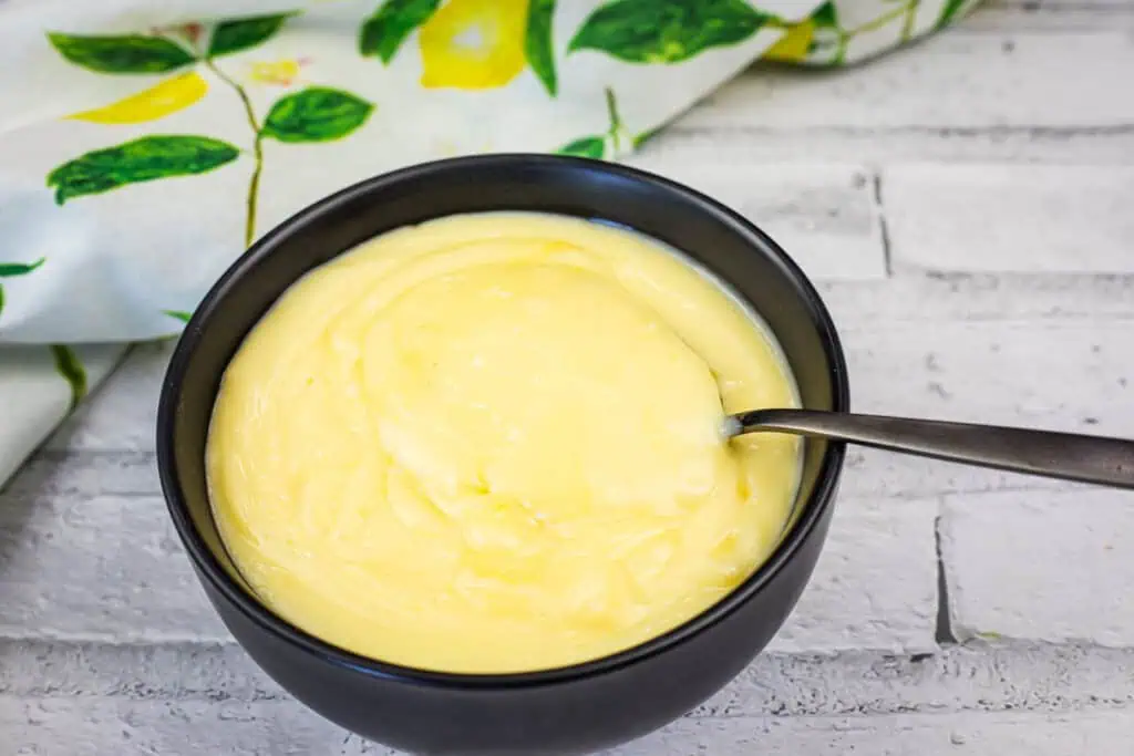 A black bowl filled with creamy yellow custard, with a spoon resting inside, placed on a white wooden surface near a patterned cloth.