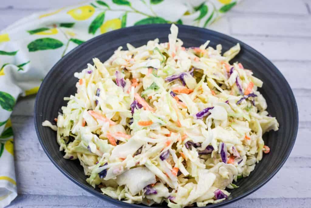 A black bowl filled with creamy coleslaw made of shredded cabbage, carrots, and purple cabbage, placed on a light-colored surface with a patterned cloth nearby.