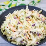 A black bowl filled with creamy coleslaw made of shredded cabbage, carrots, and purple cabbage, placed on a light-colored surface with a patterned cloth nearby.