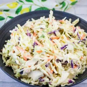 A black bowl filled with creamy coleslaw made of shredded cabbage, carrots, and purple cabbage, placed on a light-colored surface with a patterned cloth nearby.