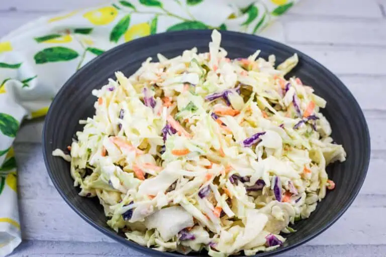 A black bowl filled with creamy coleslaw made of shredded cabbage, carrots, and purple cabbage, placed on a light-colored surface with a patterned cloth nearby.