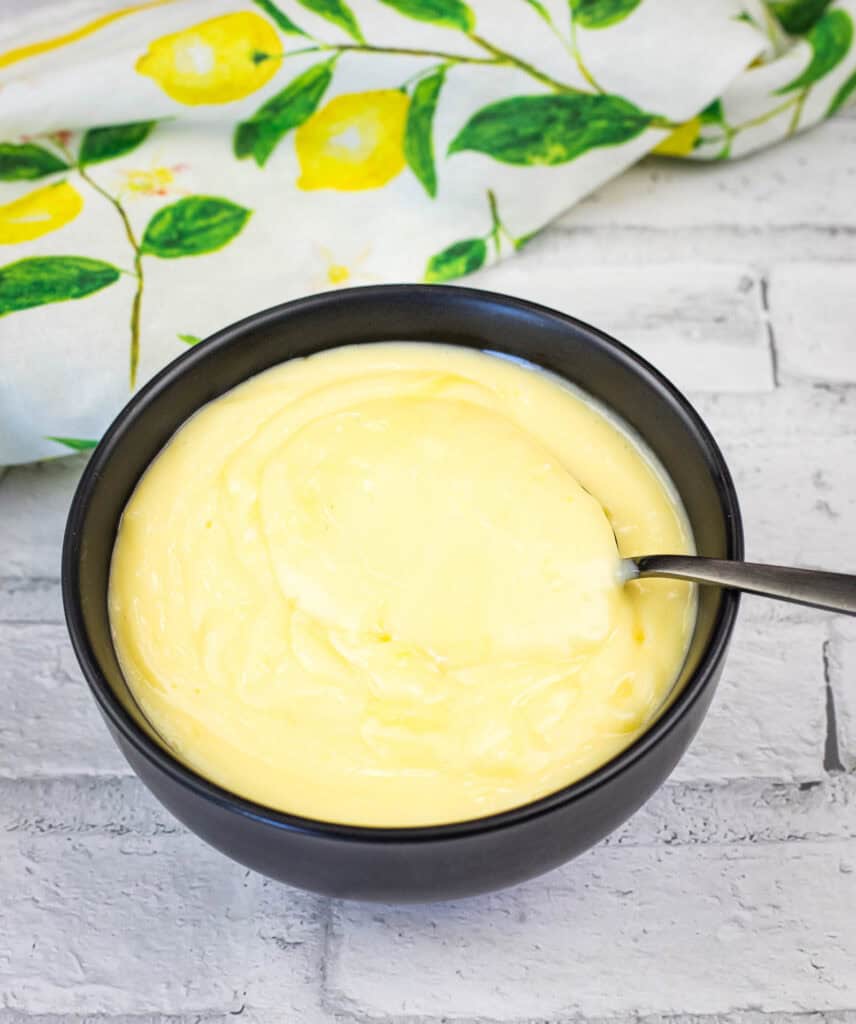 A black bowl filled with creamy yellow custard, with a spoon inside, placed on a white brick surface and a lemon-patterned cloth in the background.