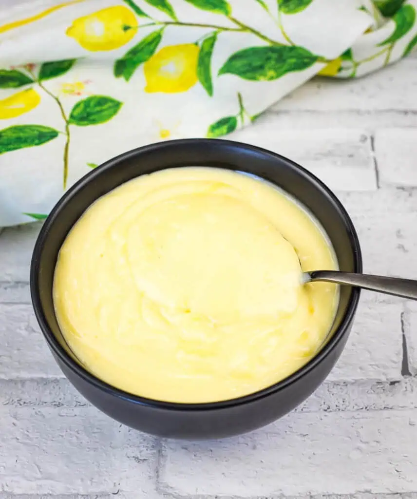 A black bowl filled with creamy yellow custard, with a spoon inside, placed on a white brick surface and a lemon-patterned cloth in the background.