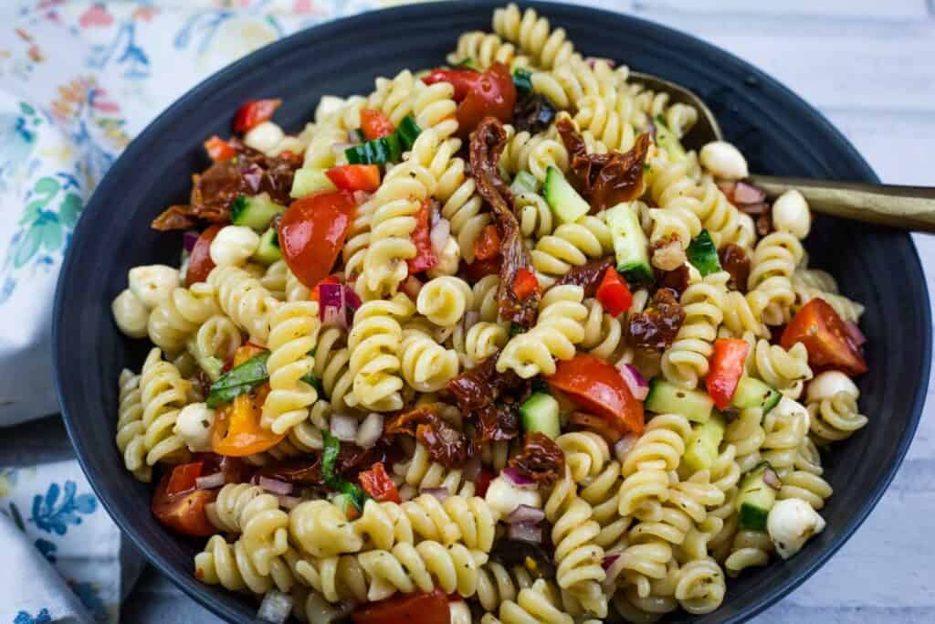 A bowl of rotini pasta salad with cherry tomatoes, cucumbers, mozzarella balls, sun-dried tomatoes, red onion, and herbs.