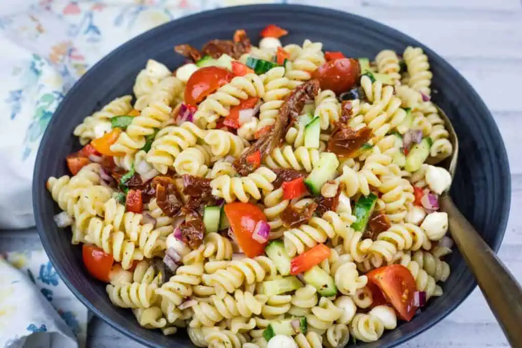 A bowl of rotini pasta salad with cherry tomatoes, cucumbers, red onions, mozzarella balls, sun-dried tomatoes, and red bell pepper.