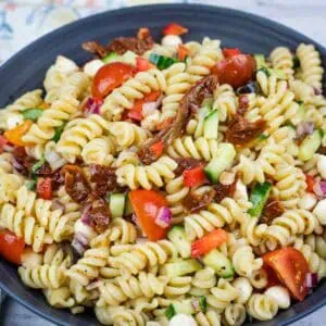 A bowl of rotini pasta salad with cherry tomatoes, cucumbers, red onions, mozzarella balls, sun-dried tomatoes, and red bell pepper.