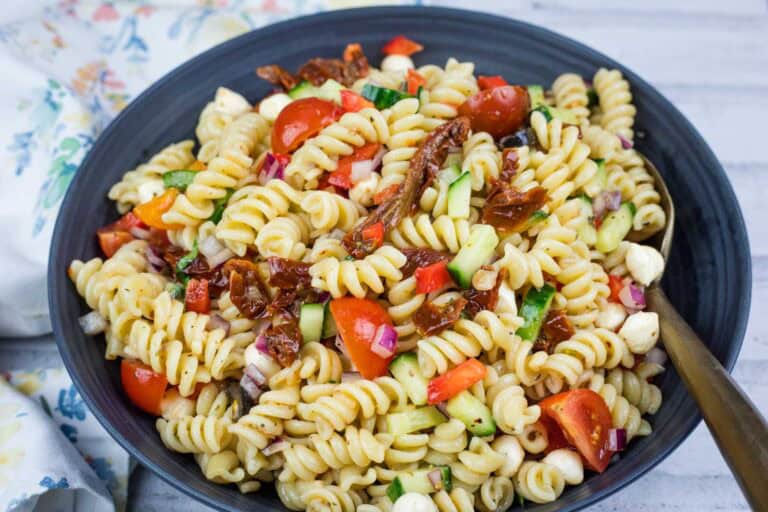 A bowl of rotini pasta salad with cherry tomatoes, cucumbers, red onions, mozzarella balls, sun-dried tomatoes, and red bell pepper.