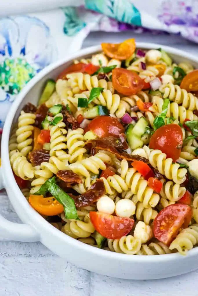A bowl of pasta salad with rotini, cherry tomatoes, mozzarella balls, cucumbers, red onions, basil, and sun-dried tomatoes.