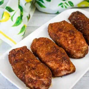 Four pieces of seasoned, cooked pork chops are arranged in a row on a white rectangular plate with a lemon-patterned cloth in the background.