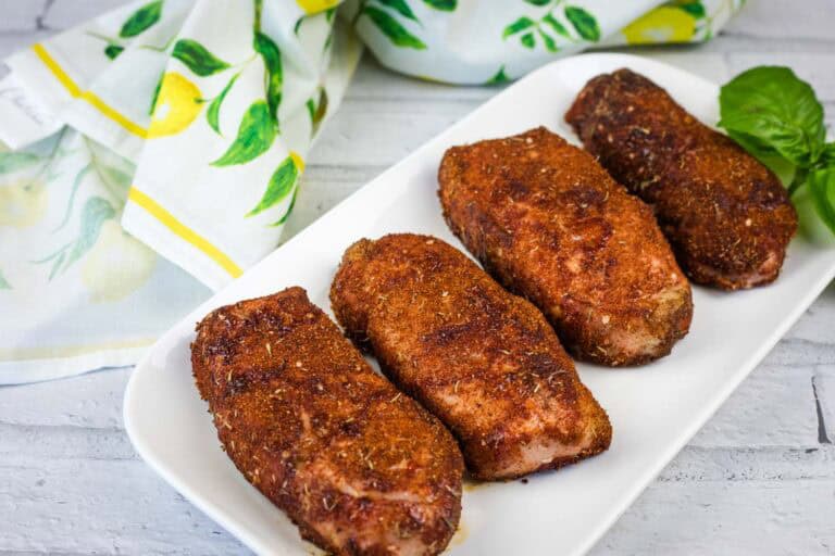 Four pieces of seasoned, cooked pork chops are arranged in a row on a white rectangular plate with a lemon-patterned cloth in the background.
