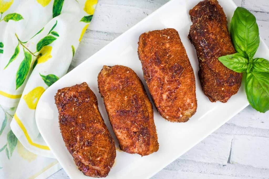 Four pieces of seasoned, cooked pork chops are arranged in a row on a white rectangular plate next to a sprig of fresh basil.