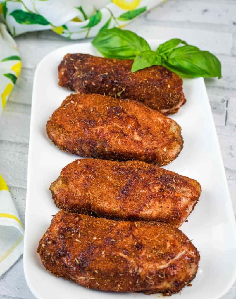 A white rectangular plate with four breaded and fried croquettes, garnished with fresh basil leaves, on a light wooden surface.