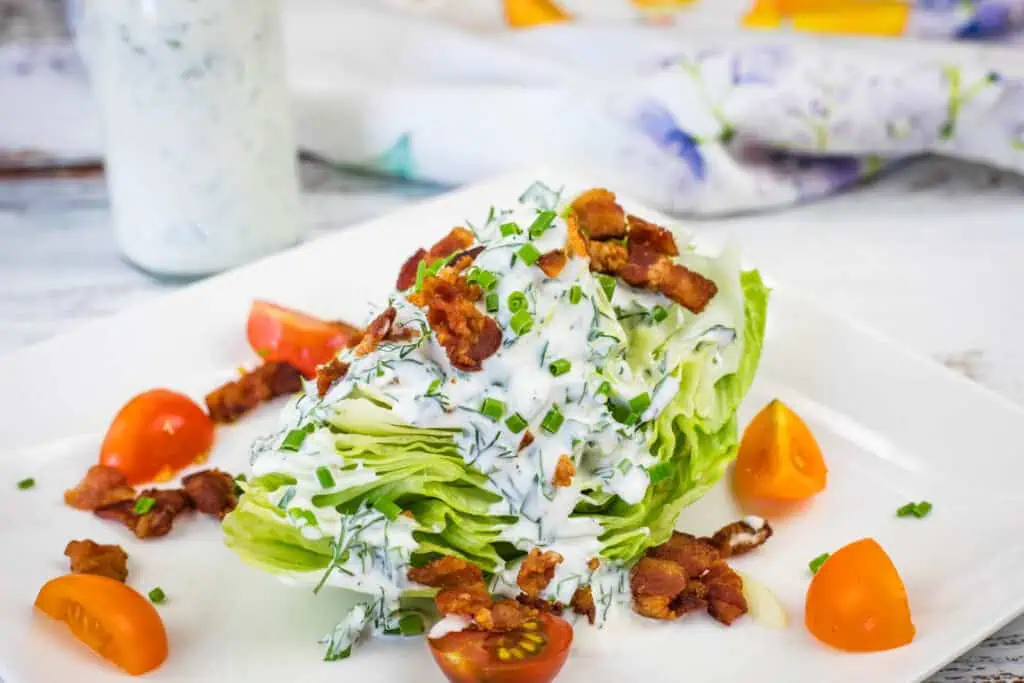 A wedge of iceberg lettuce topped with creamy dressing, bacon bits, and chopped chives, served with cherry tomato halves on a white plate.