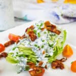 A wedge of iceberg lettuce topped with creamy dressing, bacon bits, and chopped chives, served with cherry tomato halves on a white plate.