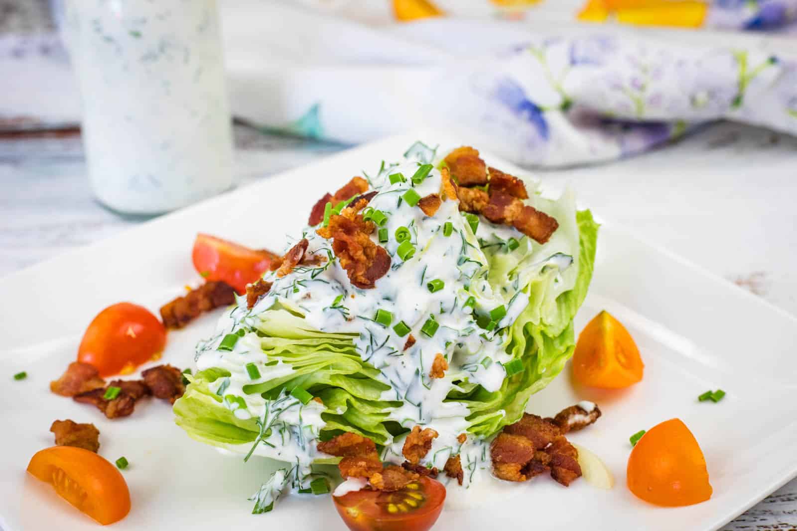 A wedge of iceberg lettuce topped with creamy dressing, bacon bits, and chopped chives, served with cherry tomato halves on a white plate.