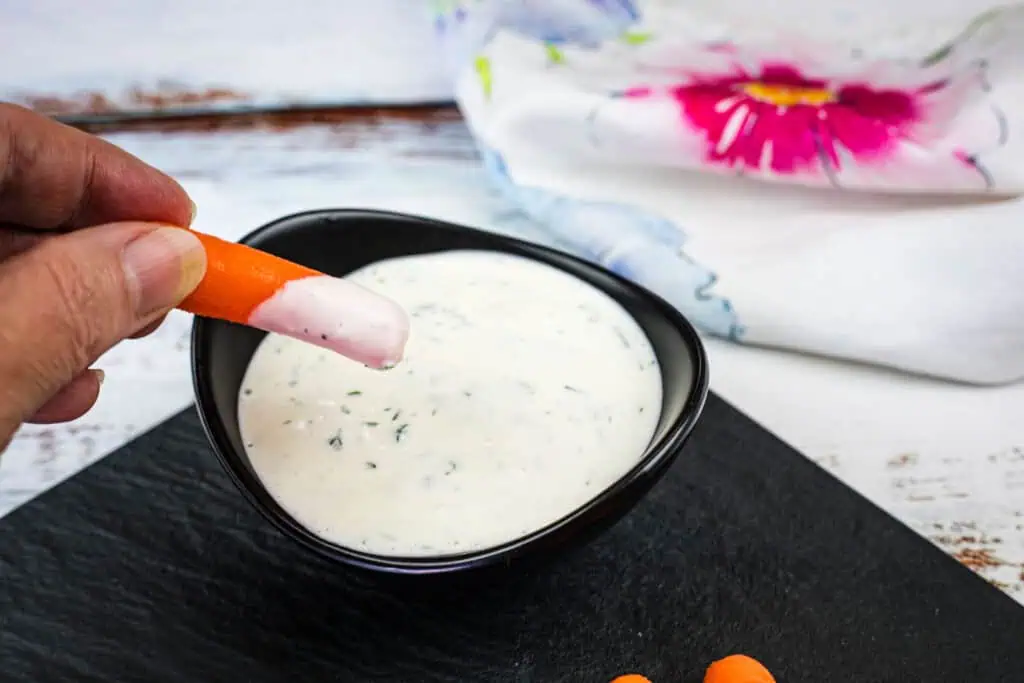 A hand dips a baby carrot into a bowl of creamy white ranch dressing on a black tray, with a floral napkin in the background.