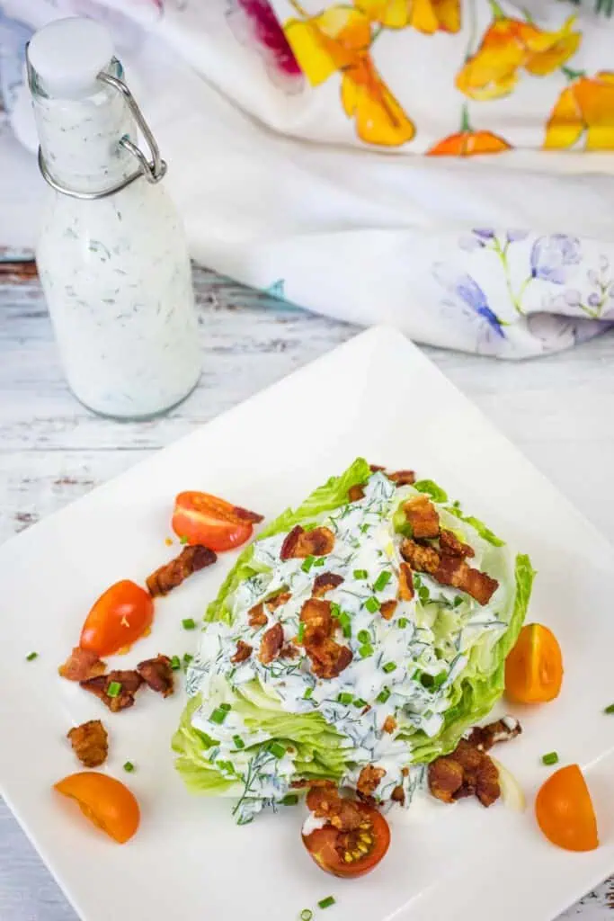 A wedge salad topped with creamy dressing, chopped bacon, chives, and tomato wedges on a square white plate, with a bottle of dressing and a floral napkin in the background.