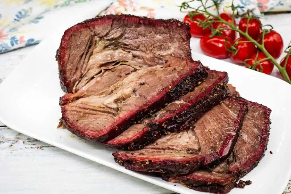 Sliced smoked beef brisket arranged on a white plate with a vine of cherry tomatoes in the background.