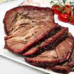 Sliced smoked beef brisket arranged on a white plate with a vine of cherry tomatoes in the background.