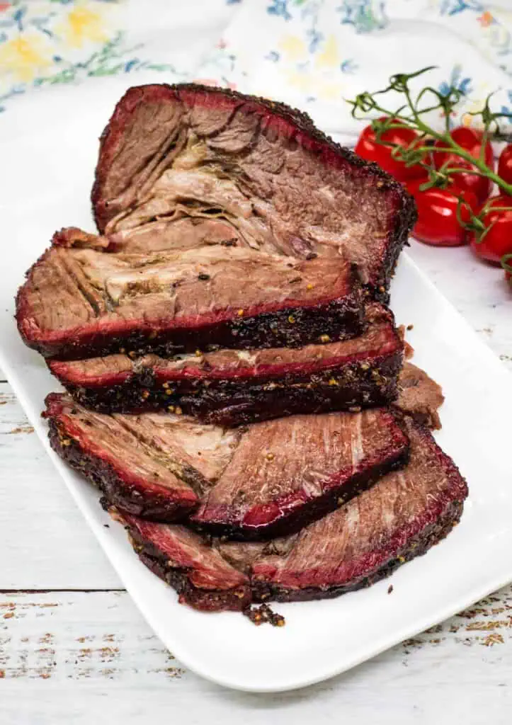 Sliced smoked brisket arranged on a white rectangular plate, with a cluster of vine tomatoes in the background.