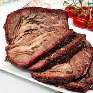 Sliced smoked beef brisket arranged on a white plate with a vine of cherry tomatoes in the background.