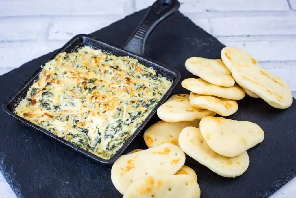 A skillet of baked spinach and cheese dip is served on a slate board with several pieces of mini naan bread arranged beside it.