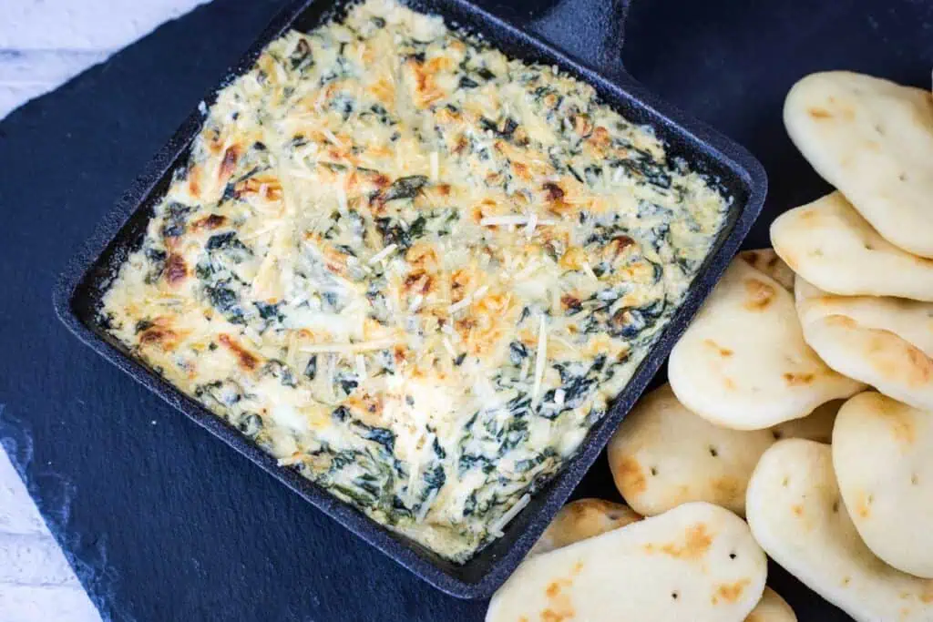 A square black dish filled with baked spinach artichoke dip, topped with melted cheese, is served next to several pieces of flatbread.