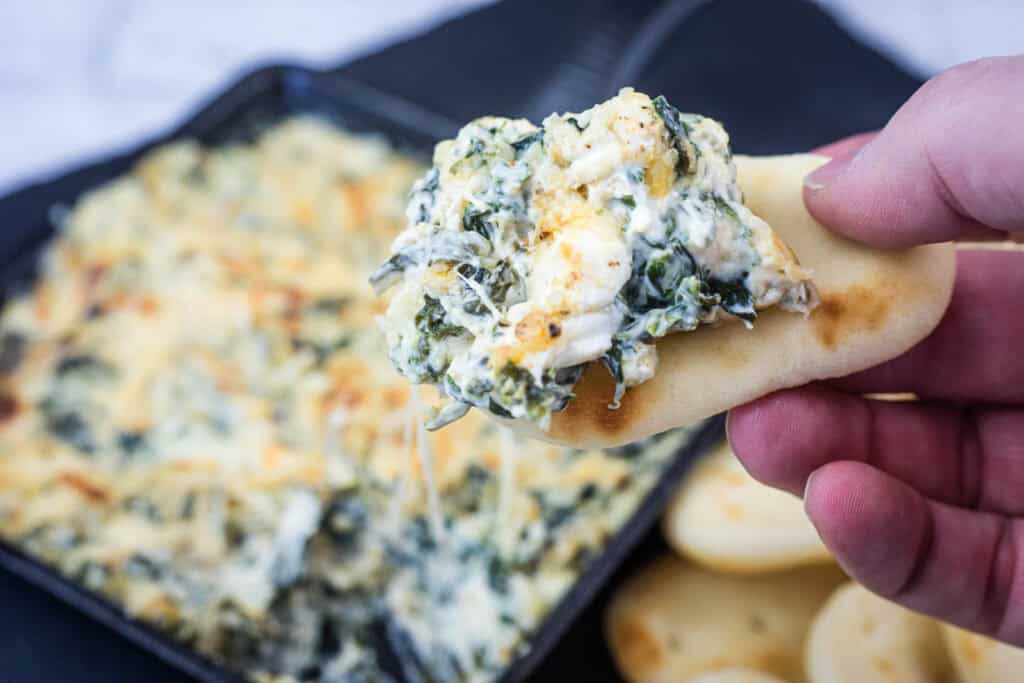 A hand holds a piece of bread topped with creamy spinach and cheese dip, with a dish of the dip and more bread pieces in the background.
