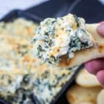 A hand holds a piece of bread topped with creamy spinach and cheese dip, with a dish of the dip and more bread pieces in the background.