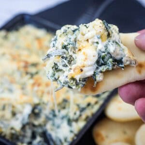 A hand holds a piece of bread topped with creamy spinach and cheese dip, with a dish of the dip and more bread pieces in the background.