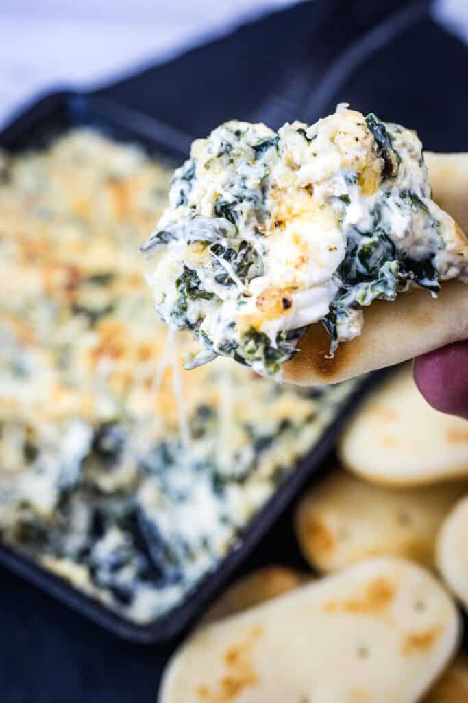 A hand holds a piece of bread topped with creamy spinach and cheese dip, with more dip and slices of bread visible in the background.