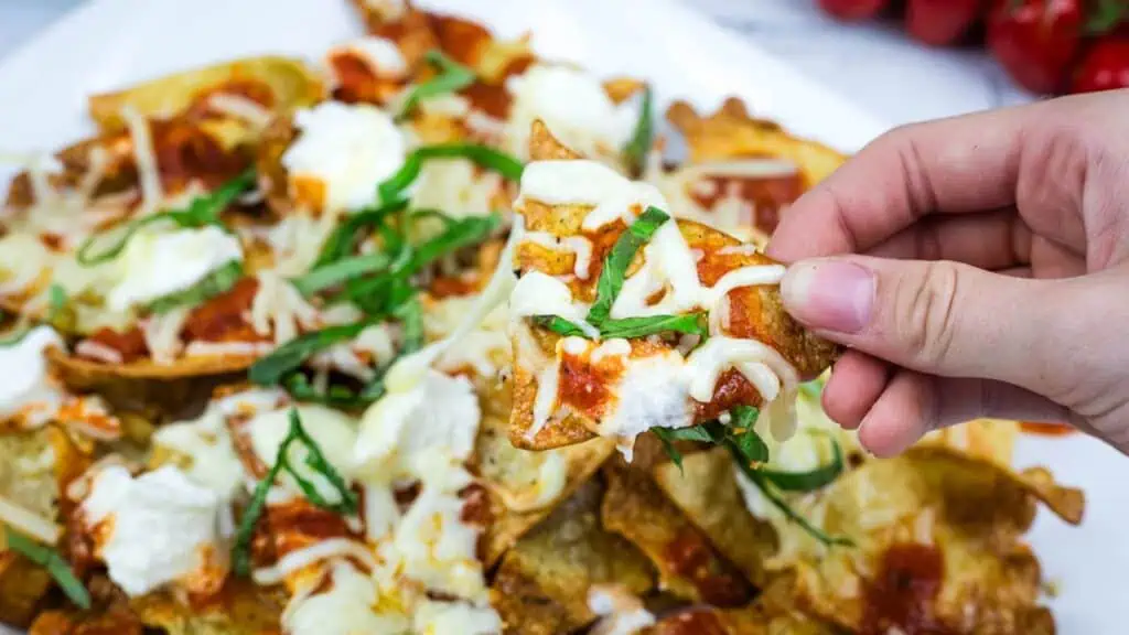 A hand holds a tortilla chip topped with melted cheese, tomato sauce, ricotta, and fresh basil over a plate of similarly topped nachos.