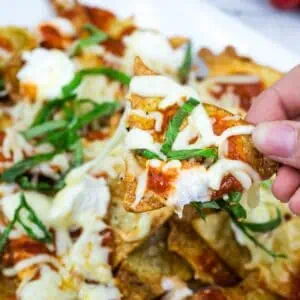 A hand holds a tortilla chip topped with melted cheese, tomato sauce, ricotta, and fresh basil over a plate of similarly topped nachos.
