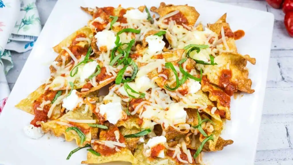 A plate of nachos topped with melted cheese, tomato sauce, ricotta, and sliced basil on a white square dish.