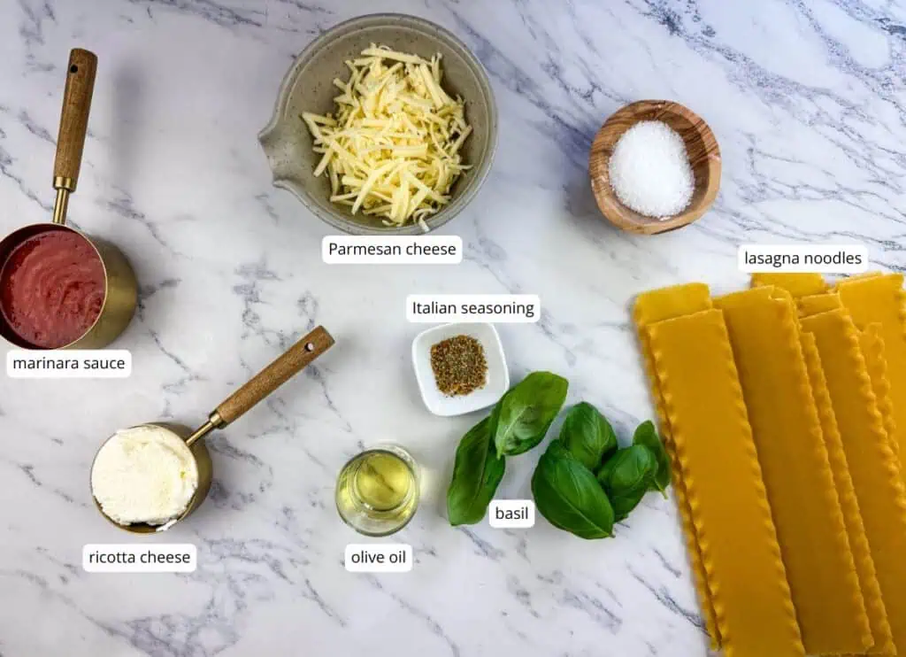 Ingredients for lasagna are arranged on a marble surface: marinara sauce, ricotta cheese, parmesan cheese, salt, Italian seasoning, olive oil, basil, and uncooked lasagna noodles.
