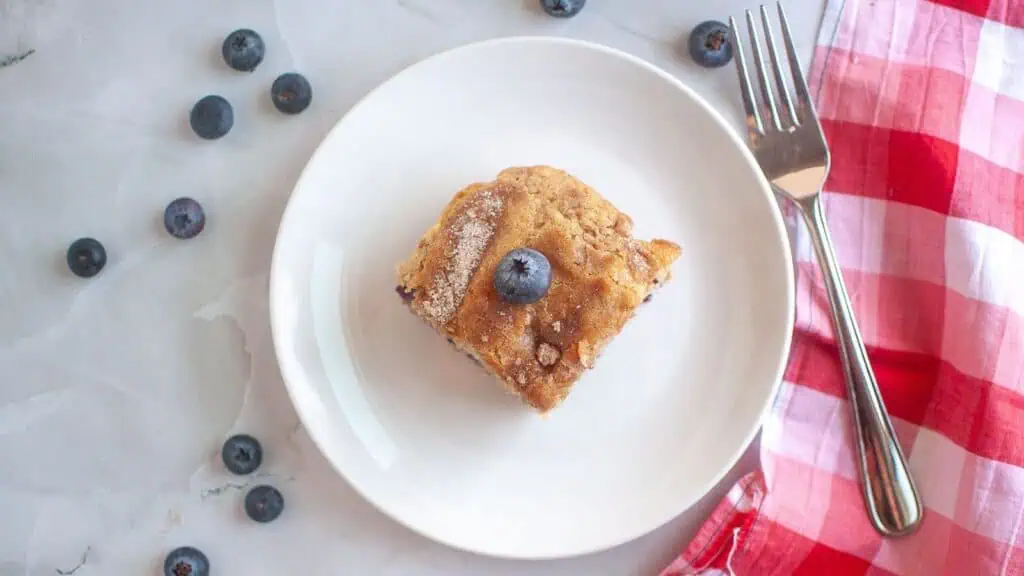 A square piece of blueberry cake on a white plate, garnished with a blueberry, with scattered blueberries, a fork, and a red-and-white napkin beside it.
