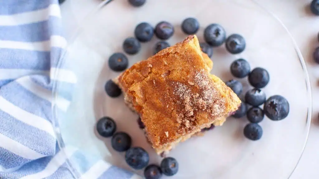 A square piece of blueberry crumb cake sits on a clear plate, surrounded by fresh blueberries, next to a blue and white striped cloth.