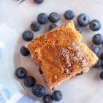 A square piece of blueberry boy bait sits on a clear plate, surrounded by fresh blueberries, next to a blue and white striped cloth.