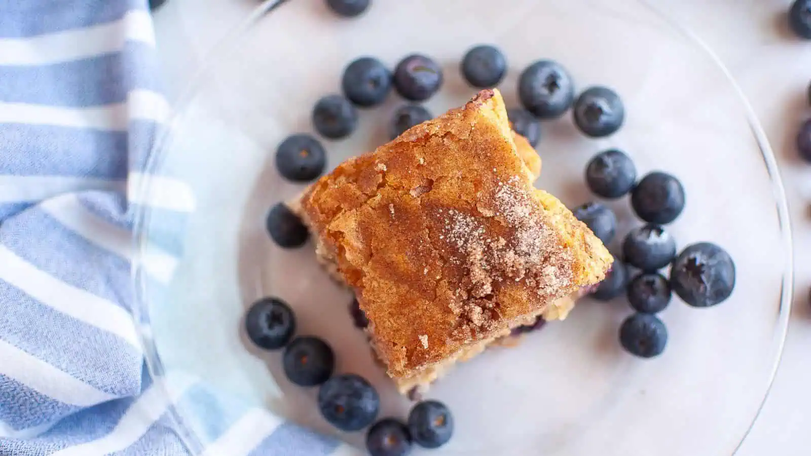 A square piece of blueberry boy bait sits on a clear plate, surrounded by fresh blueberries, next to a blue and white striped cloth.