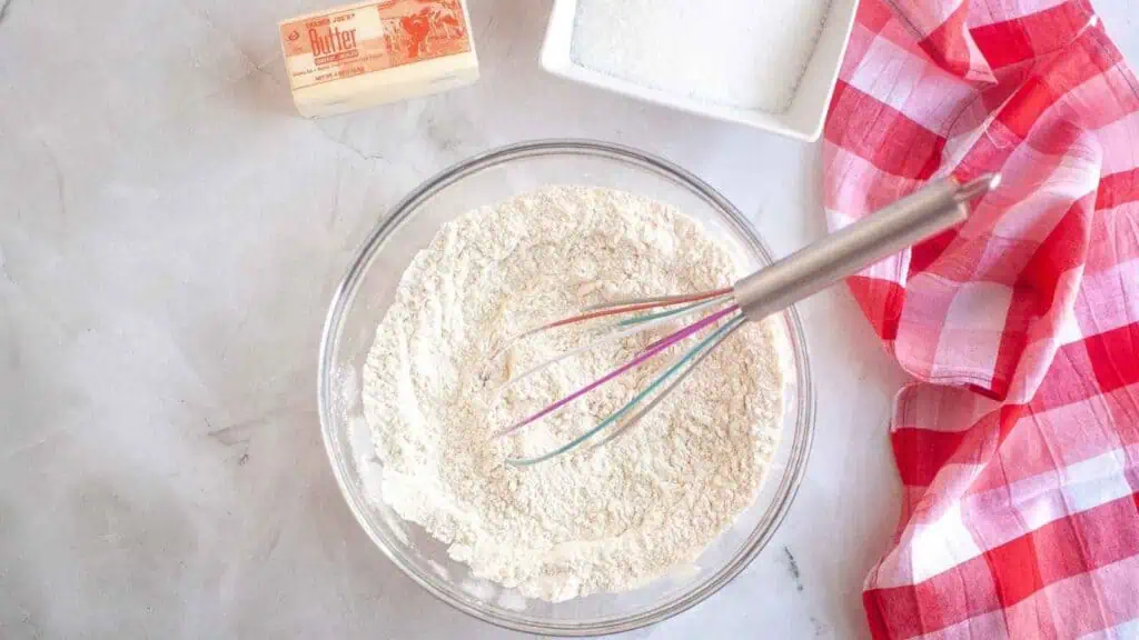 A glass bowl with flour mixture and whisk, a stick of butter, a bowl of sugar, and a red checkered cloth on a white surface.