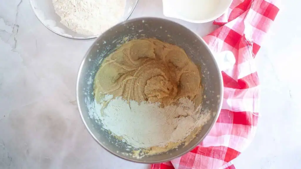 A mixing bowl with partially combined batter and flour, surrounded by a glass bowl of flour, a white bowl, and a red checkered cloth on a marble countertop.