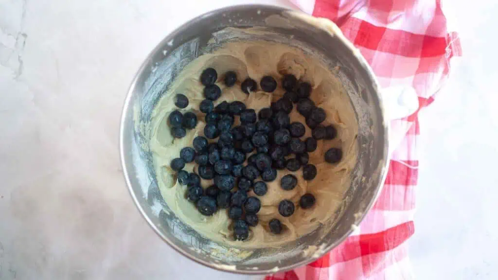 A mixing bowl with cookie dough and fresh blueberries sits on a red and white checkered cloth.