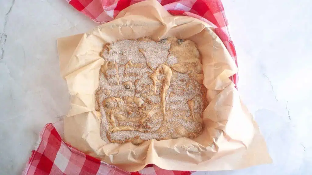 Unbaked dough covered in cinnamon sugar sits in a parchment-lined baking pan on a marble surface with a red and white checkered cloth.