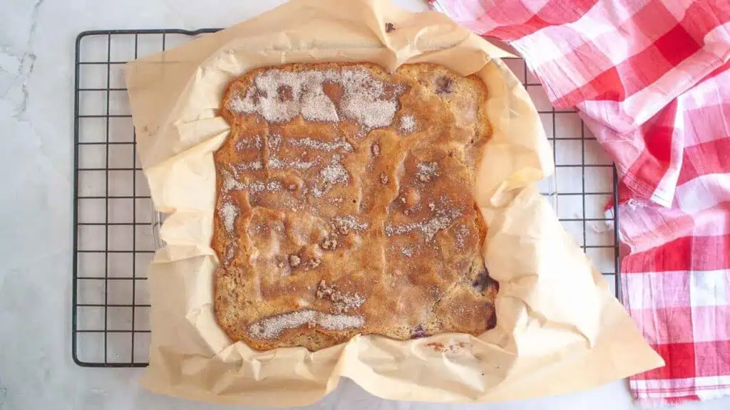 A square baked dessert topped with sugar sits on parchment paper in a baking pan, resting on a cooling rack next to a red and white checkered cloth.