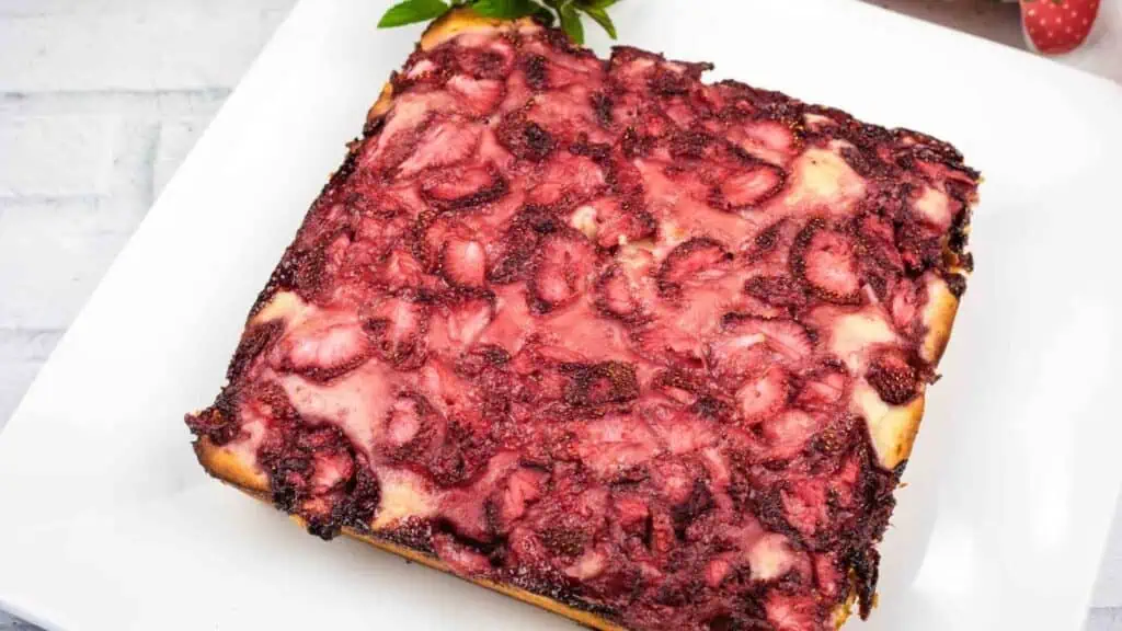 A square-shaped baked dessert with a golden crust, topped with a layer of baked strawberries, on a white plate.