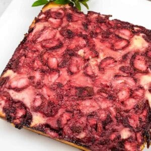 A square-shaped baked dessert with a golden crust, topped with a layer of baked strawberries, on a white plate.
