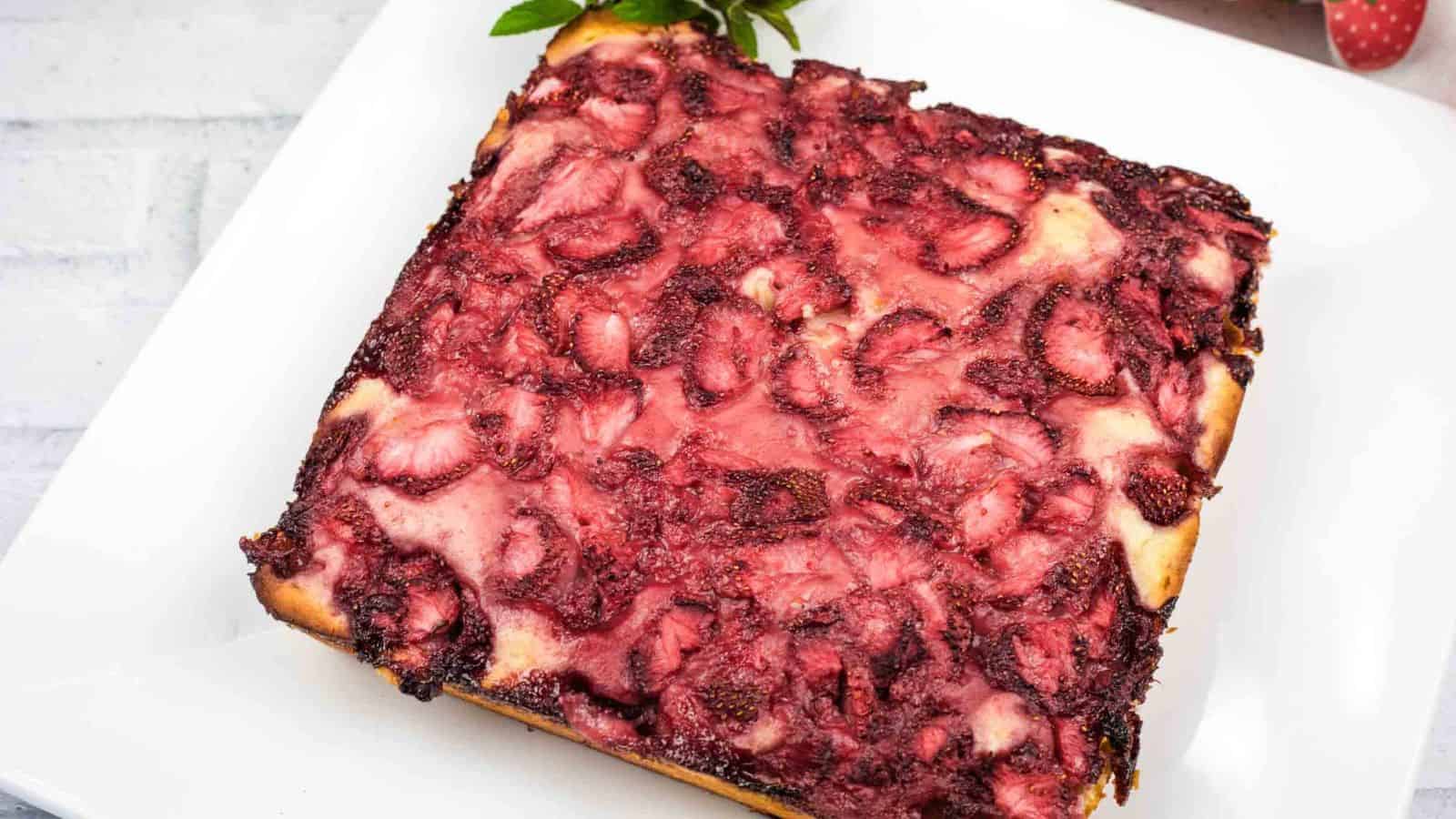 A square-shaped baked dessert with a golden crust, topped with a layer of baked strawberries, on a white plate.