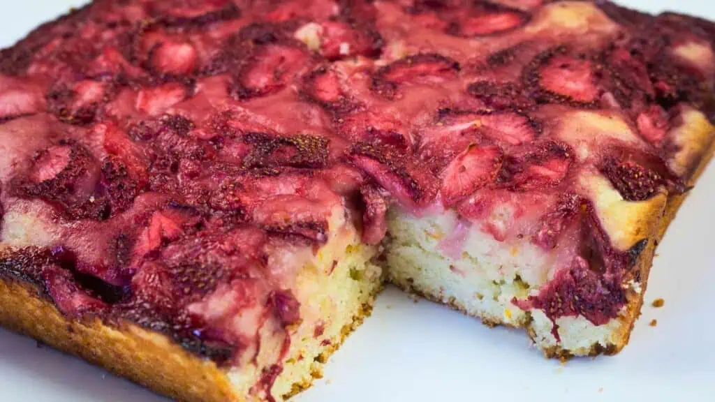 A rectangular piece of cake topped with baked strawberries has a slice cut out, revealing a light, fluffy interior.