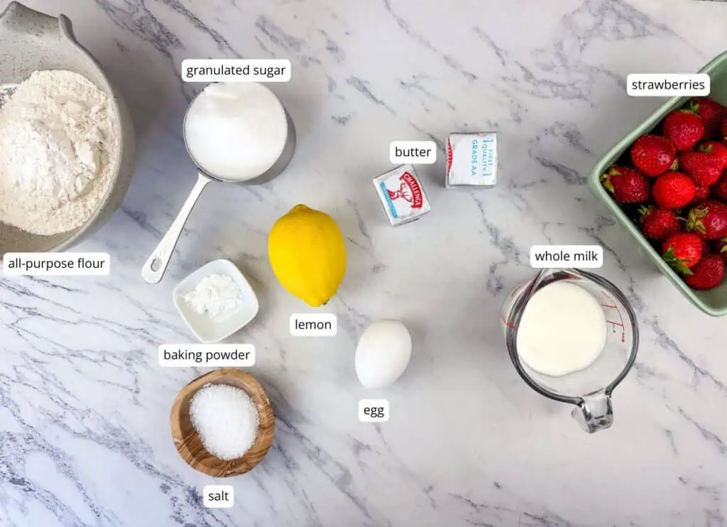Baking ingredients on a marble surface: strawberries, all-purpose flour, granulated sugar, butter, lemon, whole milk, egg, salt, and baking powder, each labeled.